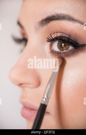 Makrodetail der braunen Mandelaugen des kaukasischen Modells während der Make-up-Sitzung. Der Make-up-Künstler wendet mit dem Pinsel einen braunen Lidschatten an. Das Modell Stockfoto