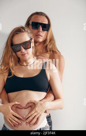 Ehemann hält den Bauch seiner Schwangeren, trägt eine Sonnenbrille und macht Symbol-Herzhände. Eine Schwangere und ein liebevoller, gutaussehender Mann umarmen zu Hause den Bauch Stockfoto