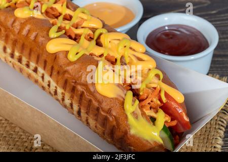 Hot Dog mit Wurst in einer Papierbox mit Tomaten und Mais, Chilischoten, Gurken und Salat. Auf einem roten Hintergrund. Fastfood. Kopierplatz Stockfoto