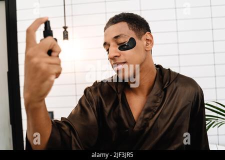 Schwarzer Mann mit Augenflecken, der Gesichtsnebel im Badezimmer des Hotels aufsprüht Stockfoto