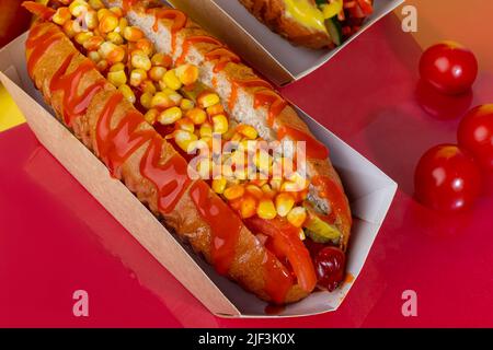 Hot Dog mit Wurst in einer Papierbox mit Tomaten und Mais, Chilischoten, Gurken und Salat. Auf einem roten Hintergrund. Fastfood. Kopierplatz Stockfoto