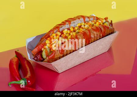 Hot Dog mit Wurst in einer Papierbox mit Tomaten und Mais, Chilischoten, Gurken und Salat. Auf einem roten Hintergrund. Fastfood. Kopierplatz Stockfoto