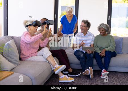 Multirassische Senioren mit digitalem pc, die Freunde betrachten, die den Virtual-Reality-Simulator auf dem Sofa genießen Stockfoto