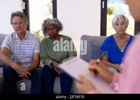 Multirassische Senioren, die weibliche Therapeutin beim Schreiben auf Papier in einer Gruppentherapie betrachten Stockfoto