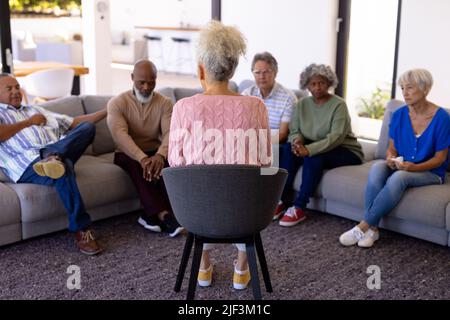 Multirassische Therapeutin im Gespräch mit Senioren, die in der Gruppentherapie im Pflegeheim auf dem Sofa sitzen Stockfoto