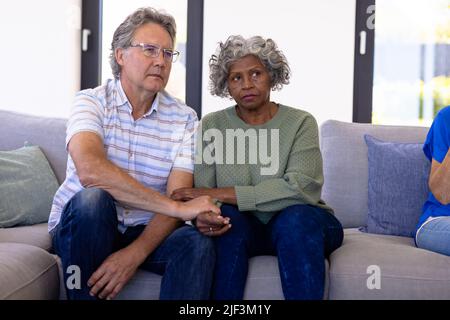 Ältere Freunde aus verschiedenen Rassen halten sich die Hände und hören in der Gruppentherapie zu, während sie auf dem Sofa sitzen Stockfoto