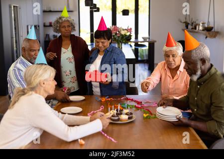 Multirassische ältere Freunde, die Partyhüte tragen, bereiten sich vor, während eine Frau ein Geburtstagsgeschenk hält Stockfoto