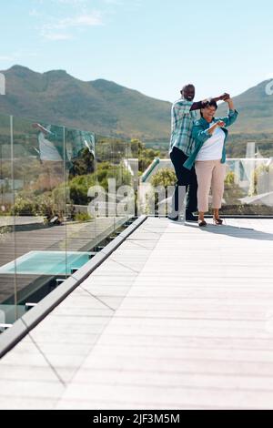 Glückliche, multirassische Senioren, die im Sommer auf dem Balkon Salsa tanzen, gegen Berg und Himmel Stockfoto