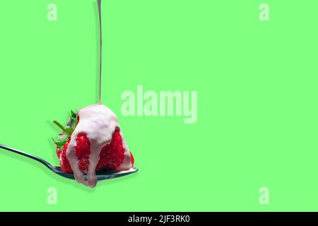 Die Creme wird auf einem Löffel über eine einzelne reife Erdbeere gegossen, auf einem farbenfrohen mintgrünen Hintergrund Stockfoto