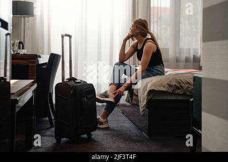 Frau, die in einem sonnigen Hotelzimmer auf dem Bett Schuhe auszieht. Touristen entspannen sich auf dem Hotelzimmer, nachdem sie mit dem Koffer unterwegs waren. Glückliches Weibchen, das Ruhe hat Stockfoto