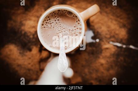 Die Milch wird in einen beigen Becher mit warmem Kakao gegossen, dessen Spritzer auf den Tisch fielen. Chaos in der Küche. Köstlicher Eiskaffee. Stockfoto