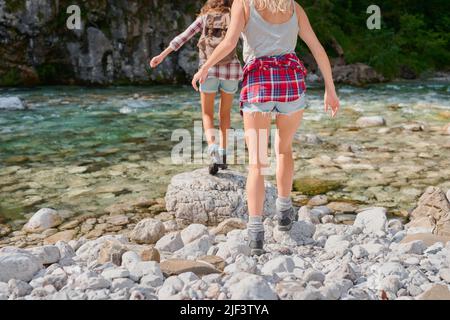 Unverkennbare Freunde, die auf einem Reise-Abenteuer gemeinsam einen Fluss überqueren und die Natur erkunden. Nahaufnahme von zwei Frauen, die über Felsen in einem See gehen, haben Stockfoto