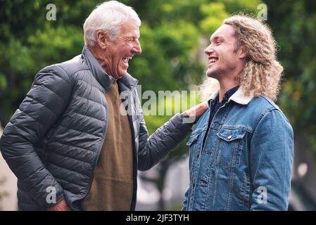 Ein reifer Mann spricht mit seinem jungen Sohn und lacht, während er seine Schulter berührt. Ein stolzer Vater, der sich mit seinem Sohn im Park lachend und sprechend entspannt Stockfoto
