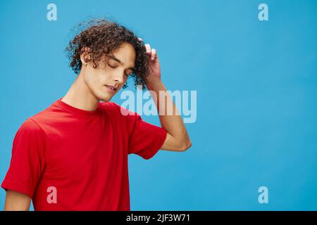 Nachdenklich nachdenklich unglücklich junge lockig gebräunt kaukasischen Kerl in grundlegenden roten T-Shirt posiert isoliert auf blauem Hintergrund. Lifestyle und Emotionen Stockfoto