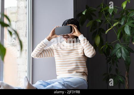 Junge erstaunt aufgeregt afro Mädchen trägt VR-Helm entspannen mit Virtual Reality auf Fensterbank zu Hause Stockfoto