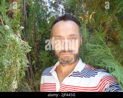 Porträt eines bärtigen Mannes mittleren Alters. Er trägt ein weißes T-Shirt. Er steht lächelnd zwischen den Bäumen. Stockfoto