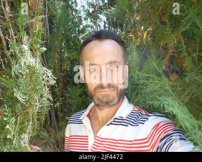 Porträt eines bärtigen Mannes mittleren Alters. Er trägt ein weißes T-Shirt. Er steht lächelnd zwischen den Bäumen. Stockfoto