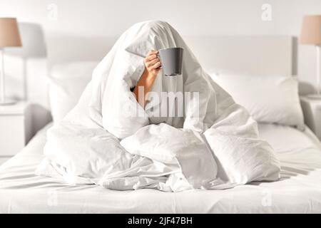 Frau mit Kaffee, die unter der Decke im Bett saß Stockfoto