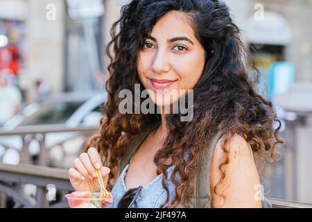 Junge dunkelhaarige Frau, die die Kamera anschaut, während sie einen Plastikbecher mit Fruchtstücken in der Hand hält Stockfoto