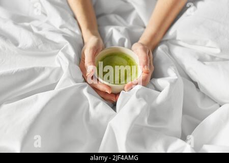 Hände einer Frau mit einer Tasse Matcha-Tee im Bett Stockfoto
