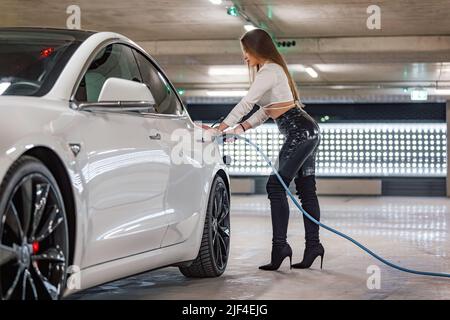 Attraktive junge Frau lädt ein Luxus-Elektroauto an einer Ladestation für Elektrofahrzeuge in einer öffentlichen Garage Stockfoto