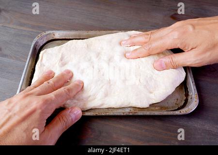 Menschenhände, die den aufgestiegenen Teig in einer Pfanne zum Backen von Pizza oder Brot strecken Stockfoto