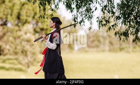 Junge asiatische Frau in traditionellen Kimono-Zügen in einer Kampfhaltung Nahaufnahme Porträt mit Katana Schwert Samurai Kriegerin Mädchen im grünen Sommergarten Stockfoto