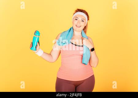 Fröhliche junge Plus-Size-Frau mit sportlichem Outfit mit einer Flasche Wasser nach dem Training auf gelbem Studiohintergrund stehend. Stockfoto