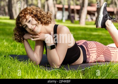 Fröhliche Rothaarige Frau in einem schwarzen Sport-BH, die auf dem Stadtpark steht, auf einer Yogamatte im Freien liegt und mit süßem Ausdruck auf die Kamera schaut. Im Freien Stockfoto