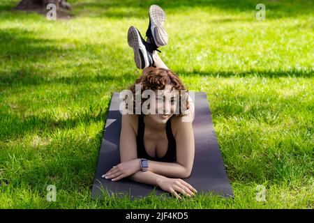 Junge Rotschopf-Frau in einem schwarzen Sport-BH, die auf dem Stadtpark steht, auf einer Yogamatte im Freien liegt und mit süßem Ausdruck auf die Kamera schaut. Im Freien Stockfoto