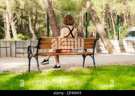 Schöne Rothaarige Frau in einem schwarzen Sport-BH, die auf dem Stadtpark steht, draußen auf einer Bank sitzt und zuschaut. Zeit allein verbringen. Denken Sie darüber nach Stockfoto