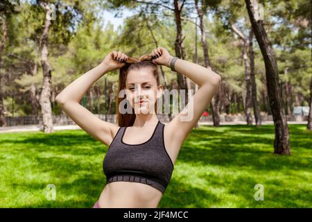 Seitenansicht einer jungen Brünette in sportlicher Kleidung im Stadtpark, im Freien, die ihre Haare in einem Pferdeschwanz bindet, während sie auf die Kamera schaut. Stockfoto