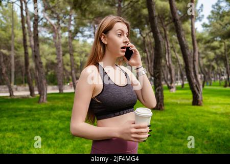 Junge, schöne Frau, die im Stadtpark sportliche Kleidung trägt, im Freien telefonieren und Kaffeebecher zum Mitnehmen hält, während sie überrascht wegschaut Stockfoto