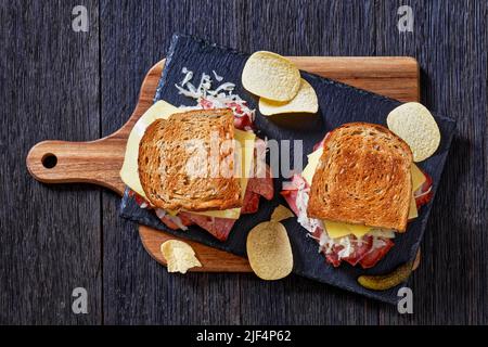 amerikanisches reuben-Sandwich mit Roggenbrot in dünne Scheiben geschnittenes Corned Beef, Sauerkraut, russischem Dressing und geschmolzenem Käse an Bord mit Kartoffelchips im Dunkeln Stockfoto