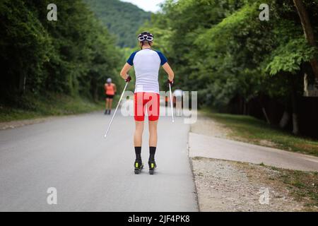 Eine Gruppe von Athleten trainiert vor dem Wettkampf. Blick von hinten. Biathlon-Fahrt auf den Rollskiern mit Skistöcken, im Helm. Konzept des Sports Stockfoto