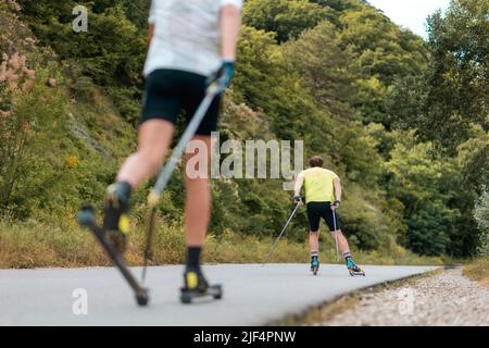 Biathlon-Workout. Zwei sportliche Männer trainieren auf dem Rollski auf der Landstraße, Rückansicht. Low-Angle-Ansicht. Konzept des Sportwettbewerbs und des Sommers ACT Stockfoto