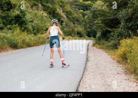 Athletische junge Frau, die auf dem Rollski auf der Landstraße trainiert, Rückansicht. Speicherplatz kopieren. Konzept von Wettbewerb, Biathlon und Sommertraining. Stockfoto