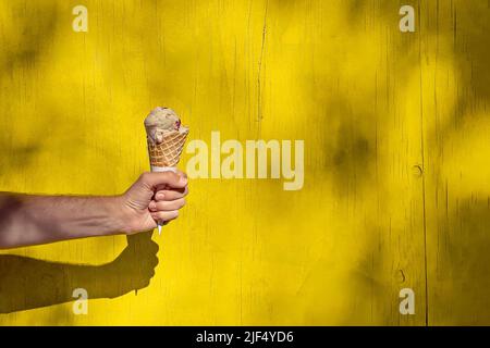 Die Hand des Mannes hält an einem sonnigen Sommertag ein Eis im Kegel über der hellgelben Wand. Natürliche Schatten, Kopierbereich Stockfoto