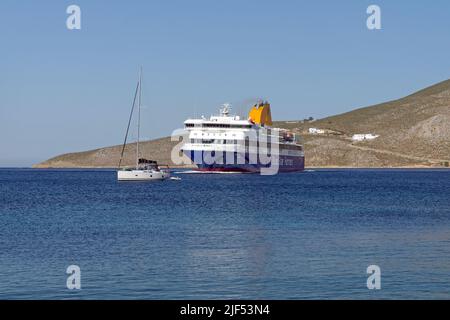 Blue Star, die Patmos Fähre, manövriert um ein kleines Segelboot, als sie zum Hafen von Livadia, Tilos Insel, in der Nähe von Rhodos anlegt. Mai 2022 Stockfoto