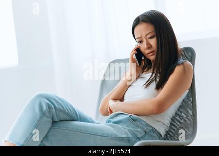 Verwirrt aufgeregt gebräunt schöne junge asiatische Frau hören schockiert Nachrichten beim Anruf mit Telefon zu Hause innen Wohnzimmer. Fernkommunikation Social Stockfoto