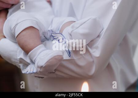 Taufe von Säuglingen. Nahaufnahme von winzigen Babybeinen, Sakrament der Taufe. Der Pate hält das Kind in den Armen. Stockfoto