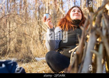 Frau mit rothaarigen Haaren entspannt sich am sonnigen Wintertag in Lotusposition bei einer Tasse Tee in erhobener Hand und genießt die Natur. Stockfoto