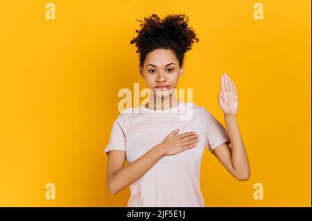 Nur die Wahrheit. Ehrliche, stolze, lockige, junge afroamerikanische Frau im lässigen T-Shirt, die Hand auf der Brust hält und die Handfläche hebt, Versprechen gibt, Eid ablegt, auf isoliertem orangefarbenem Hintergrund steht Stockfoto