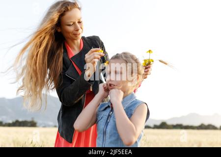 Mutter flechtet die Haare der Tochter mit gelben Blüten auf dem Feld. Stockfoto