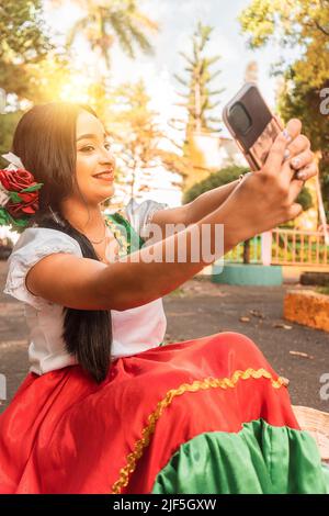 Schöner lateinischer Teenager in traditioneller Kleidung, der an einem sonnigen Tag ein Selfie in einem Park gemacht hat Stockfoto