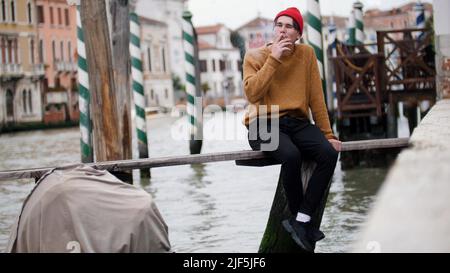 Ein junger Mann sitzt auf der Bank über dem Wasser und raucht Stockfoto
