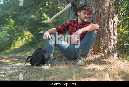 Holzfällerarbeiter, der im Wald sitzt. Holzfäller mit Axt auf Waldgrund. Mann, der seine Arbeit verführt. Holzfäller, der im Wald steht Stockfoto