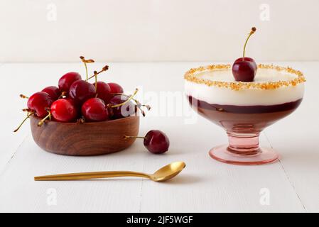 Dunkle und weiße Schokoladenmousse oder Panna Cotta mit Kirschgelee in einem Glas mit Holzschüssel mit reifen Kirschen auf weißem Holztisch. Weitwinkelansicht, Stockfoto