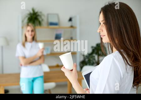 Tolle Zeit beim Mittagessen. Zwei freundliche Praktikanten in weißer Uniform trinken Kaffee im Büro und lächeln. Der Arbeitsplatz der Docs ist so modern und Stockfoto