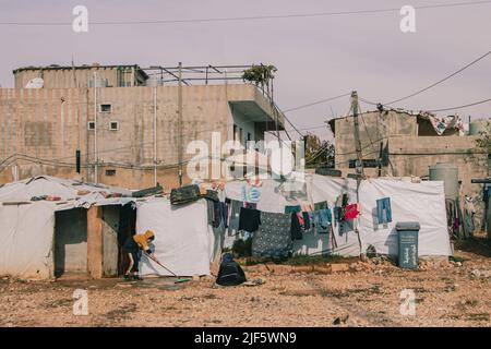 Ein Mädchen putzt den Boden vor ihrem Zelt, während ihre Mutter auf dem Boden betet. An einem Morgen in einem Flüchtlingslager im Bekaa-Tal leben rund 20 Familien. Einige Familien warten seit fünf Jahren darauf, an einen anderen Ort zu gehen. Selbst wenn die Situation ihnen eine Rückkehr nach Syrien ermöglichen würde, sind die meisten von ihnen nur canít. Die im Libanon geborenen Kinder haben keinen syrischen Pass, sind aber im Land registriert. Deshalb bleiben auch die Eltern auf dem Land. Viele Eltern haben auch ihre Aufenthaltserlaubnis abgelaufen und können es sich nicht leisten, eine neue auszustellen, da viele von zwei bis drei Dollar leben Stockfoto